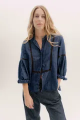 Camisa de jean azul oscuro con cuello mao, botones frontales, mangas largas abullonadas con puños elásticos y detalles bordados en color marrón en los hombros y mangas.