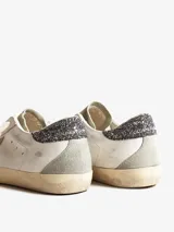 Championes Golden Goose modelo Super-Star, confeccionados en cuero color blanco con paneles de gamuza gris. Presentan el icónico parche de estrella lateral, detalle de glitter en el talón, efecto envejecido en la suela y logo GGDB grabado en el lateral.
