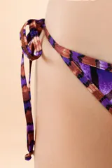 Bombacha de bikini con estampado de hojas en tonos púrpura y cobre, con tiritas laterales ajustables y ribetes decorativos.