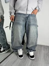 Jeans baggy de denim con efecto acid wash y lavado desgastado, de corte holgado. Presenta amplios bolsillos traseros con bordados contrastantes, incluyendo un diseño geométrico en rojo y blanco en un bolsillo y un motivo de querubín con la inscripción "Ameri" en el otro.