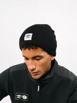 Gorro tipo beanie de tejido acanalado en color negro, con parche rectangular blanco con el logo de Umbro bordado en el frente.