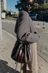 Bolso tipo saca de cuero sintético color marrón oscuro, con diseño fruncido y cierre mediante cordón ajustable. Cuenta con asas de hombro combinadas con cadena metálica y bolsillo interior con cremallera.