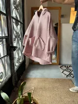 Campera con capucha confeccionada en tejido tipo corderito color rosa, con diseño de collage geométrico en el frente que incluye bloques de color en naranja, negro y blanco. Presenta mangas largas de corte holgado y diseño artesanal único.