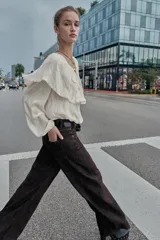 Blusa de color beige con escote en pico y lazo frontal decorado con abalorios. Presenta mangas largas con puños elásticos y un detalle de volantes amplios con terminación de puntilla a tono sobre los hombros y el pecho.