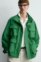 Chaqueta tipo cazadora de corte relajado en color verde, confeccionada en tejido de mezcla de algodón. Presenta cuello subido con forro interior a cuadros, manga larga con puños abotonados y cuatro bolsillos frontales de plastrón con solapa. Incluye cierre frontal de cremallera oculta por solapa con botones a presión y cintura ajustable en el interior.