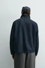 Chaqueta de denim de algodón en color azul oscuro, con cuello solapa y manga larga. Presenta cierre frontal con cremallera oculta bajo una solapa con alamares metálicos, bolsillo de vivo en el pecho y bolsillos de plastrón con solapa en la cadera.