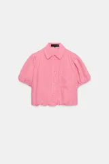 Camisa corta de cuello solapa y manga corta abullonada acabada en elástico, color verde con estampado de cuadros vichy. Cuenta con un bolsillo de plastrón en el delantero, bajo con elástico y cierre frontal con botones.