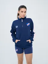 Buzo de felpa azul marino con medio cierre, cuello alto, bandas laterales en tono rosado y bolsillo frontal tipo canguro. Presenta el logo de Umbro y el escudo del Club Nacional de Football bordados.
