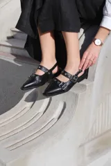 Zapato de cuero negro con punta fina, dos tiras con hebillas plateadas y detalle de ojales metálicos.