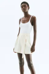 Short de tiro alto color blanco roto, con pinzas frontales y un cinturón fijo con anillas metálicas decorativas en un lateral.