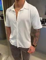 Camisa de manga corta color blanco, de corte clásico y tela liviana con sutil textura de líneas. Cuenta con cuello camisero y cierre frontal de botones.