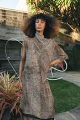 Blusa de cuello subido y manga sisa, confeccionada en tejido fluido semitransparente con estampado de lunares en tonos beige y negro. Presenta un diseño superpuesto tipo capa con caída asimétrica y cierre posterior con abertura y botón.
