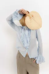 Camisa de estilo romántico confeccionada en algodón, con cuello alto, volados frontales y mangas largas con puños acampanados también decorados con volados.