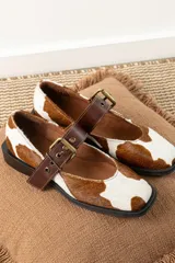 Zapatos tipo Mary Jane con punta cuadrada, confeccionados en cuero con estampado de vaca (animal print) y tira ajustable con hebilla metálica en color marrón.