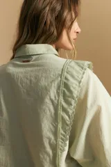 Camisa blanca de manga larga con cuello y botones frontales. Presenta detalles de volados o puntilla en los hombros y mangas abullonadas con puños anchos. La tela parece tener un sutil tejido a rayas verticales.
