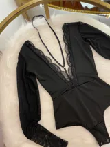 Body de lycra y encaje color negro, con mangas largas de tul transparente y detalle de encaje en los puños. Presenta un escote profundo con detalle de encaje y un collarín integrado que se conecta al centro del pecho.