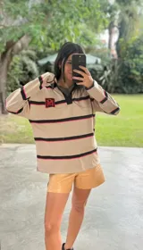 Buzo tipo rugby de algodón con diseño de franjas horizontales en tonos beige, negro y fucsia. Presenta cuello polo en contraste y cierre frontal parcial.