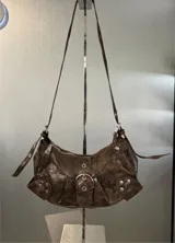 Cartera tipo hobo de cuero sintético con efecto desgastado, color marrón. Presenta una hebilla central decorativa, detalles de tachas metálicas en los laterales y correa ajustable.