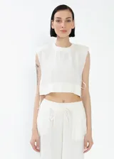 Remera crop top de lino color blanco, con cuello redondo y hombreras marcadas que le dan una estructura definida. Presenta un corte recto y holgado con aberturas laterales ajustables mediante cordones.