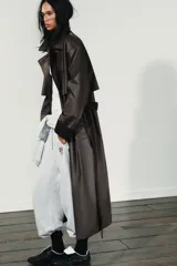 Gabardina larga de efecto encerado en color marrón oscuro, con cuello solapa y cierre frontal cruzado con botones. Presenta mangas largas con puños vueltos en pana, bolsillos laterales y cinturón ajustable de pana. Incluye pinza en la espalda.