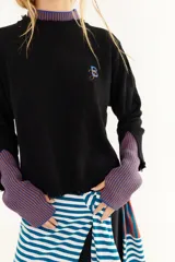 Sweater de punto color marrón con detalles en rojo en el cuello y los puños. Presenta un diseño de doble capa en el cuello y mangas largas con orificios para los pulgares, además de un pequeño bordado en el pecho y un acabado desflecado en el ruedo.