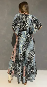 Vestido largo con escote en V tanto en el frente como en la espalda, mangas cortas amplias y corte elastizado debajo del busto. Presenta un estampado de estilo paisley y guardas geométricas en tonos azules, negros y corales.