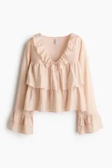 Blusa rosada de tejido liviano, con escote en V rodeado de volados y tiras finas para anudar. Presenta un diseño escalonado con volados anchos en el pecho y en el bajo, y mangas largas acampanadas con volados dobles en los puños.