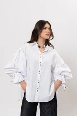 Camisa de algodón blanca de silueta amplia, con cuello camisero, botonadura frontal con botones decorativos, mangas abullonadas extragrandes y detalles de tiritas ajustables con cuentas de resina en los puños.