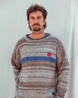 Sweater tejido a mano de calce holgado, con diseño de rayas horizontales en tonos tierra y una franja azul central. Presenta un bordado con las letras 'SM' en color rojo en la parte frontal.