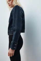 Chaqueta de denim de corte corto, color azul oscuro, con cuello redondo, hombreras y cierre frontal con cremallera. Presenta diseño de inspiración militar con doble botonadura metálica decorativa en el frente, trabillas con botón en los hombros y bajo elástico.