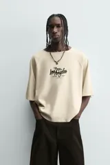 Camiseta de hombre, corte relaxed fit, color beige claro con textura. Presenta cuello redondo amplio y manga corta. Estampado frontal en negro con texto que incluye "Los Angeles".