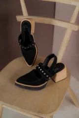 Zapatos tipo zueco de gamuza negra con punta fina y tira sobre el empeine decorada con tachas metálicas plateadas. Posee taco cuadrado de madera y suela con plataforma.