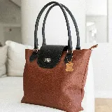 Cartera tote de cuero vacuno color marrón con solapa negra y cierre metálico. Tiene doble asa de mano negra, bolsillo interno con cierre y cierres YKK metálicos.