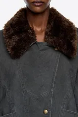 Cazadora corta de corte cruzado confeccionada en mezcla de algodón lavado color gris oscuro. Presenta un cuello camisero con pelo sintético desmontable en color marrón, cierre frontal con botones de presión y mangas largas con trabillas en los puños. Incluye bolsillos laterales, espalda fruncida en la cintura y trabillas ajustables laterales para definir la silueta.