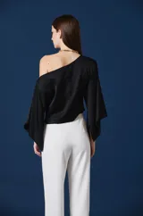 Blusa de mujer color negro, con diseño asimétrico de un solo hombro y mangas murciélago amplias. Presenta un aplique decorativo en forma de libélula dorada en el hombro derecho.