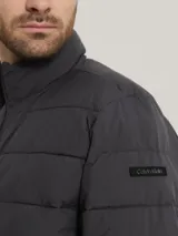 Campera puffer Calvin Klein color gris oscuro, con cuello alto, cierre frontal con cremallera, bolsillos laterales con cierre, puños elásticos y logo en la manga.