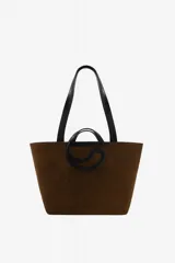 Bolso tipo tote amplio de ante marrón, con doble asa larga negra para el hombro y detalle gráfico frontal en forma de C con asas cortas integradas.
