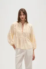 Blusa de viscosa color crema con detalles de bordado y broderie. Presenta un diseño holgado, cuello en V con cierre de botones y mangas abullonadas con puños elásticos.