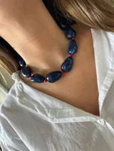 Collar corto con cuentas grandes e irregulares de color azul y pequeñas cuentas rojas intercaladas. Posee cadena de alargue y herrajes de acero inoxidable.