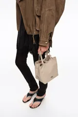 Bolso shopper mediano de nylon color beige con estructura blanda, doble asa corta de mano y correa fina ajustable. Cuenta con cierre de botón a presión, bolsillo interior con cierre y aplique metálico con el logo de la marca en el frente.