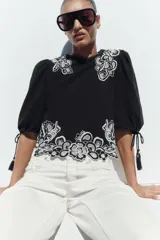 Blusa corta negra con mangas abullonadas y bordados florales en contraste de color blanco. Presenta cuello redondo con puntilla, mangas ajustables con cordones y borlas, y cierre en la espalda con abertura y botón oculto.