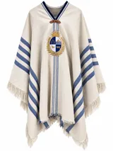 Poncho de hilo de algodón grueso color beige, con detalles de rayas azules y flecos en los bordes. Presenta el escudo nacional uruguayo bordado en el pecho.