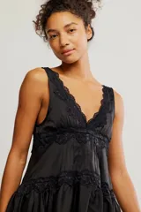 Mini vestido tipo slip de tela satinada negra, con escote en V profundo, diseño en capas con volados y detalles de encaje en los bordes. Corte estilo babydoll, sin mangas y silueta amplia.