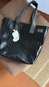 Bolso tipo tote bag negro con textura símil cuero de cocodrilo. Tiene doble asa corta, cierre superior y un dije colgante con forma de ala.
