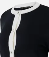 Cardigan corto de punto en color blanco con detalles en negro, cuello redondo, manga larga y cierre frontal de botones perlados con ribete negro. Cuenta con dos bolsillos delanteros de ojal con ribete negro y botón.