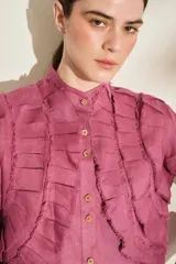 Camisa de lino color morado, con cuello mao, cierre frontal con botones y mangas largas con puños estructurados. Presenta detalles de volados frontales y ruedo con terminaciones desflecadas.