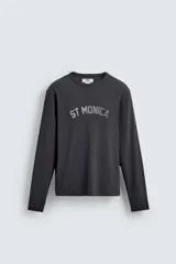 Camiseta de manga larga, corte ajustado (muscle fit), color gris oscuro con efecto lavado. Presenta un estampado frontal desgastado con el texto curvo "ST MONICA" en color plateado o gris claro.