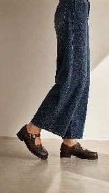 Mocasín de cuero color marrón con acabado texturizado, diseño de punta redonda con costuras artesanales visibles y cierre mediante una tira con hebilla lateral.