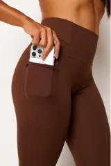 Calza deportiva de tiro alto color marrón, confeccionada en tejido técnico de alto rendimiento con compresión. Presenta bolsillos laterales funcionales y detalles reflectivos para mayor visibilidad.