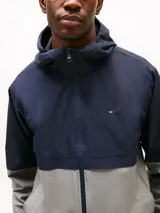 Campera deportiva Tommy Hilfiger para hombre, confeccionada en nylon, con diseño de bloques de colores azul marino y gris, capucha ajustable, cierre frontal con cremallera y logo bordado en el pecho.